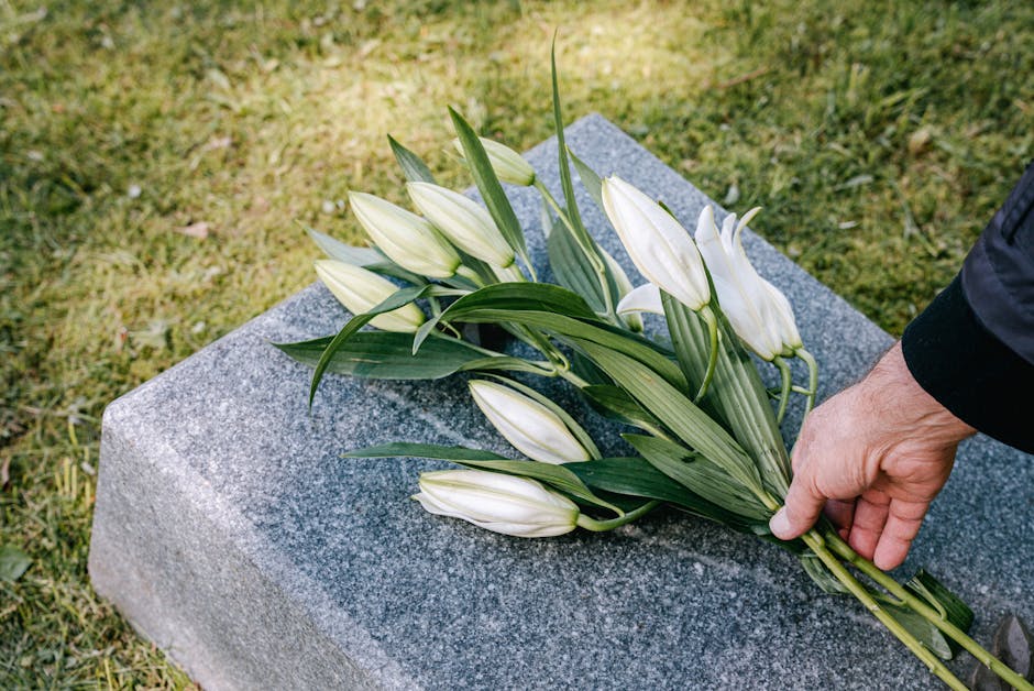 A hand carefully places a bouquet of white lilies with long, slender green leaves onto a grey granite memorial stone outdoors, with grass visible in the background. The lilies are fresh, with closed b