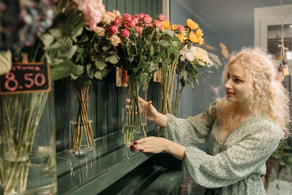 A young woman with curly blonde hair and fair skin selecting a floral arrangement at a flower shop. She is wearing a light green, floral-patterned blouse with long, billowing sleeves. She is holding a