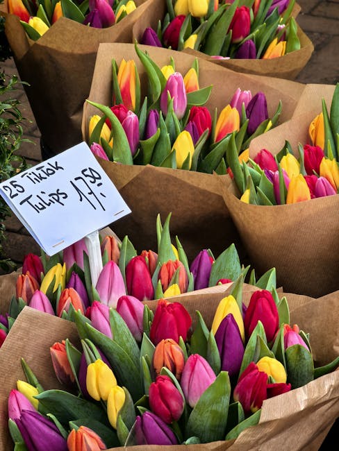 Multiple bouquets of fresh tulips in vibrant shades of pink, purple, yellow, and orange are arranged in brown craft paper wrapping on a display table. The tulips have sturdy green stems and smooth, sl