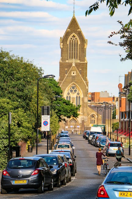 A street scene in Blackheath featuring a tall, historic church with pointed arches and stained glass windows, surrounded by lush green trees and a row of parked cars along the curb. The sky is partly 
